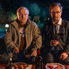 Zwei Männer sitzen mit Essen und Getränken auf einem Sofa im Freien. Ein Mann hält ein Glas Rotwein, während der andere ernst schaut.