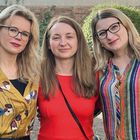 Drei lächelnde Frauen stehen zusammen im Freien. Die linke trägt eine gelbe Bluse mit bunten Blumenprints, die rechte gestreift.