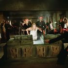 Menschen in roten Stirnbändern führen eine Frau in Weiß zu einem steinernen Altar mit Reliefs, umgeben von Fackeln.