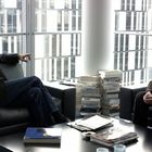 Zwei Männer sitzen sich in einem modernen Büro gegenüber. Im Hintergrund sind Bücherstapel und große Fenster zu sehen.