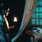 Eine Person hält eine Kerze, beleuchtet einen Mann in blutbefleckter Kleidung auf einem Bett in einem dunklen Raum.