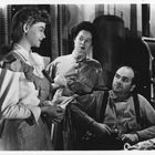 Drei Personen in einem Raum; eine Frau im karierten Kleid hält ein Tuch, während ein Mann sitzend in die Ferne blickt.