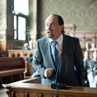 Ein Mann in einem grauen Anzug spricht leidenschaftlich in einem klassischen Gerichtssaal voller Menschen.