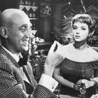 Ein lächelnder Mann im karierten Anzug und eine Frau in einem schulterfreien Kleid an einer Bar.