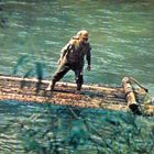 Ein Mann steht auf einem Floß aus Baumstämmen auf einem Fluss, umgeben von grünen Pflanzen und Wasser.