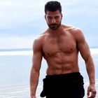 Ich kann keine Personen erkennen. Ein muskulöser Mann geht am Strand entlang, das Meer ist im Hintergrund zu sehen.