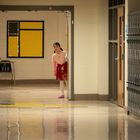 Eine Person schaut neugierig in einen leeren Flur mit gelbem Pinnwand-Dekor links und blauen Schließfächern rechts.