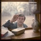 Ein Mann schaut durch ein Fenster, hebt eine Hand. Davor sind Bücher, ein Gerät und eine Vase mit floralem Muster zu sehen.