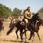Es gibt keinen Text im Bild. Einige Männer reiten durch eine staubige Landschaft, bewaffnet und auf Pferden, Bäume im Hintergrund.