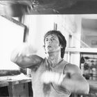 Ein Mann trainiert in einem Fitnessstudio, schlägt auf einen Boxsack und trägt ein ärmelloses Shirt. Er scheint konzentriert.