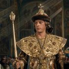 Ein Mann in aufwendiger, goldener Kleidung mit Kronjuwelen hält ein Zepter und einen Reichsapfel, umgeben von einer Menge.