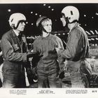 Text: "BRIGITTE BARDOT in PLEASE, NOT NOW." Drei Personen in Overalls und Helmen stehen unter Lichtern und sprechen.