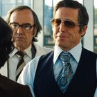 Zwei Männer in Anzügen stehen in einem hellen Raum, einer trägt eine große Brille, der andere eine gemusterte Krawatte.