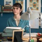 Bücherregal im Hintergrund. Frau mit Brille in blauer Strickjacke sitzt an einem Schreibtisch mit Schreibmaschine.