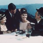 Ein Tisch mit vier fröhlichen Menschen in eleganter Kleidung, vor einer Bergkulisse. Wein und leere Gläser stehen auf dem Tisch.