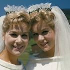 Zwei lächelnde Frauen in weißen Hochzeitskleidern und Schleiern, vor einer grünen, bergigen Landschaft.