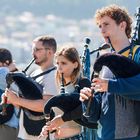 Eine Gruppe von Menschen spielt Dudelsäcke im Freien, mit Gebäuden im unscharfen Hintergrund. Sie wirken konzentriert.