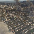 Drohnen schweben über einer großen Stadt, während dicker Rauch aus mehreren Stellen aufsteigt. Berge sind im Hintergrund sichtbar.