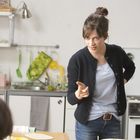 Eine Frau in einem minimalistischen, hellen Raum gestikuliert energisch in Richtung der Kamera. Küche im Hintergrund.