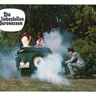 Text: "Die liebestollen Baronessen". Zwei Frauen im Auto, viel Rauch, ein Mann in Uniform hockt daneben. Grünbewuchs im Hintergrund.
