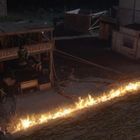 "Bear's Blacksmith" und "Post office" auf Gebäuden. Eine Flammenlinie verläuft diagonal durch eine dunkle Straßenszene.