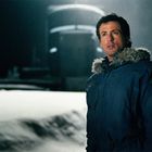 Ein Mann in einer blauen Jacke steht in einer verschneiten, dunklen Umgebung. Im Hintergrund ist ein unscharfes, großes Objekt.