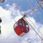 Text "8" auf einer roten Gondel. Eine Person springt von einem Riesenrad in einen blauen Himmel mit Wolken.