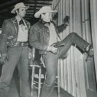 Zwei Männer in Cowboy-Kleidung, einer tritt mit dem Fuß eine Holztür ein, beide tragen Hüte und Pistolen.