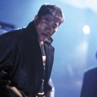 Person in dunklem Mantel, ernst blickend, steht in nebliger Nachtlandschaft mit unscharfem Hintergrund und schwachem Licht.