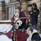 Eine Person sitzt mit einem Husky in einem Schlitten. Eine andere steht daneben und schaut auf ein Handy. Winterliche Umgebung.