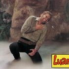 Text "Luana" in roter Schrift auf gelbem Hintergrund. Ein Mann kniet mit schmerzverzerrtem Gesicht, Hand auf der Brust, in nebliger Umgebung.