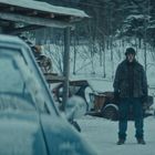 Zwei Menschen stehen sich im verschneiten Freien gegenüber. Links ein unscharfer Wagen, rechts ein Schuppen.