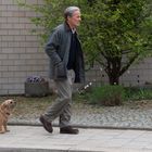 Ein älterer Mann spaziert mit einem kleinen, zotteligen Hund auf einem Gehweg vor einer Ziegelwand mit grünem Buschwerk.