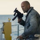 Ein Mann mit ernstem Gesichtsausdruck hält eine Waffe, Hintergrund ist das Meer. Er trägt eine Jeansjacke.