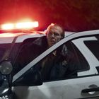 Ein Polizist steigt nachts aus einem Streifenwagen. Blaulicht strahlt, die Vorderseite des Autos zeigt die Zahl "305".