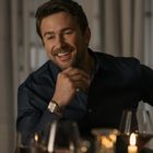 Ein Mann in dunklem Hemd lächelt, stützt sein Kinn mit der Hand. Im Vordergrund Kerzen und Weingläser.