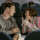 Zwei Personen in einem Flugzeug, lächelnd und sich anscheinend unterhaltend, beide halten ein Glas.