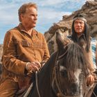 Drei Männer reiten auf Pferden durch felsige Landschaft, tragen Western- und indigene Kleidung. Der Himmel ist blau und klar.