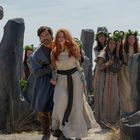 Ich sehe keinen erkennbaren Text. Eine Gruppe in traditioneller Kleidung, Kränze tragend, in einem Steinkreis. Zwei Personen im Fokus.