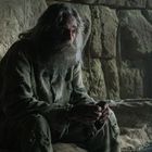 Ein älterer Mann mit langem Haar und Bart sitzt in einem kargen, steinigen Raum auf einem einfachen Bett.