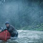 Zwei Menschen in winterlicher Kleidung ziehen ein rotes Kanu durch einen flachen, bewaldeten Fluss.