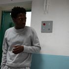 Ein Mann in grauem Pullover schaut zur Seite, in einem Raum mit blau-weiß gestrichenen Wänden; eine rote Taste an der Wand.
