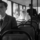 Ein junger Mann in einem Anzug sitzt nachdenklich in einem Bus. Hinter ihm sitzen eine verhüllte Frau und Männer in historischen Uniformen.