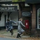 Schilder mit Texten "TAILOR SWIFT" und "FACKHAM'S FASTEST TAILOR". Zwei Menschen in grauen Mänteln rennen Hand in Hand.
