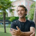 Ein lächelnder Mann in schwarzem T-Shirt steht in einem grünen Park mit Palmen; ein rosa Gebäude im Hintergrund.