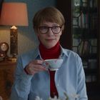 Eine Person mit Brille hält eine blumige Tasse. Im Hintergrund eine Lampe, Uhr und Bücherregal.