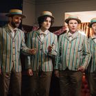 Vier Männer in gestreiften Anzügen und Strohhüten singen in einem Raum. Es ist festlich dekoriert.