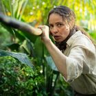 Eine Frau in tropischer Umgebung hält einen Stock und schaut konzentriert. Sie trägt ein weißes Hemd und ein Halstuch.