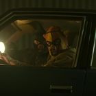 Zwei Personen in einem Auto tragen Schweinemasken. Sie blicken ernst aus dem Fenster, während eine starke Lichtquelle sie beleuchtet.