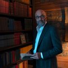 Ein Mann in einem Anzug, mit einem Buch in den Händen, steht in einer holzgetäfelten Bibliothek vor einem Regal voller Bücher.